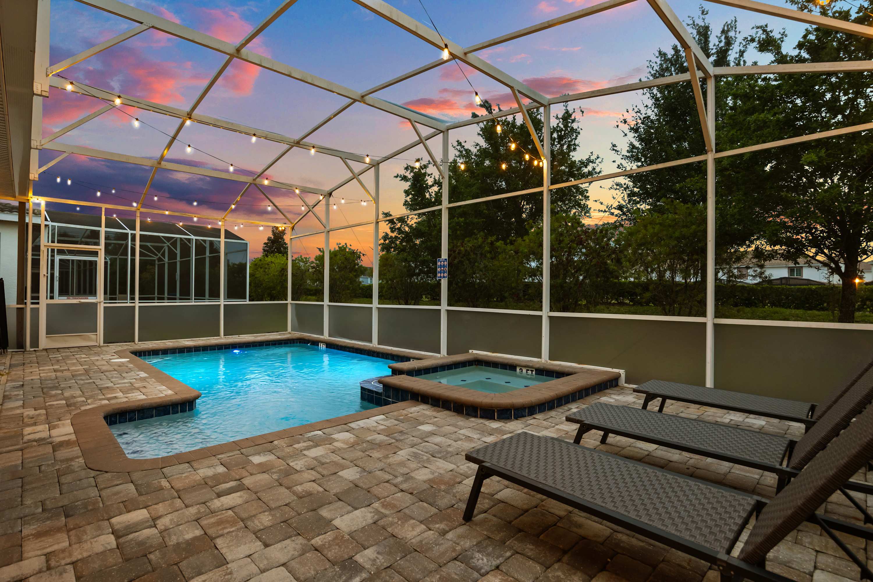 Inviting view of the private swimming pool and adjacent hot tub within a screened enclosure, surrounded by lounge chairs and illuminated by string lights under a colorful sunset.