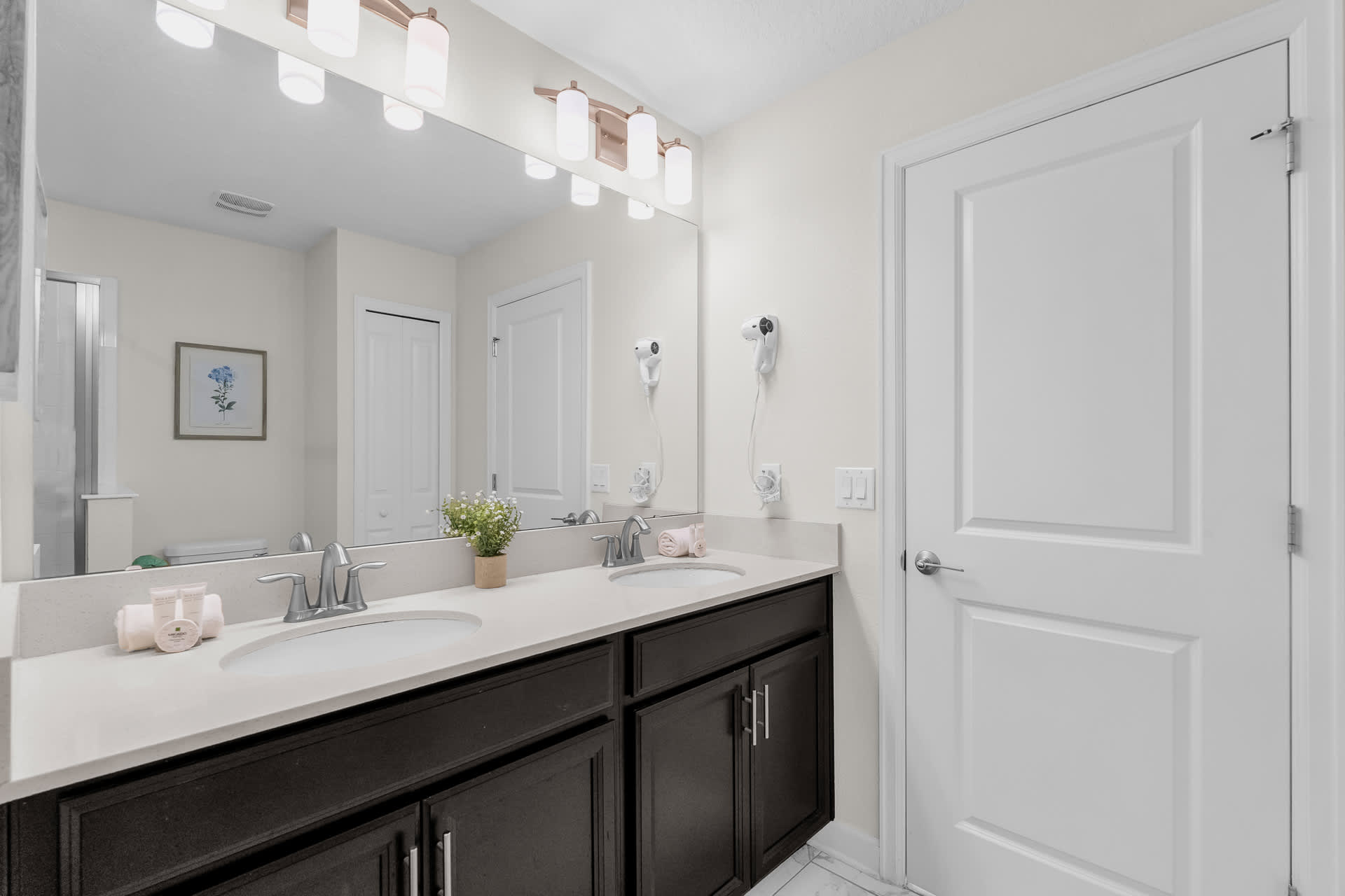 Bright bathroom featuring a double vanity with dark wood cabinets, light quartz countertops, and two rectangular sinks. The room has white tiled floors and a large mirror reflecting a white door and a closet.