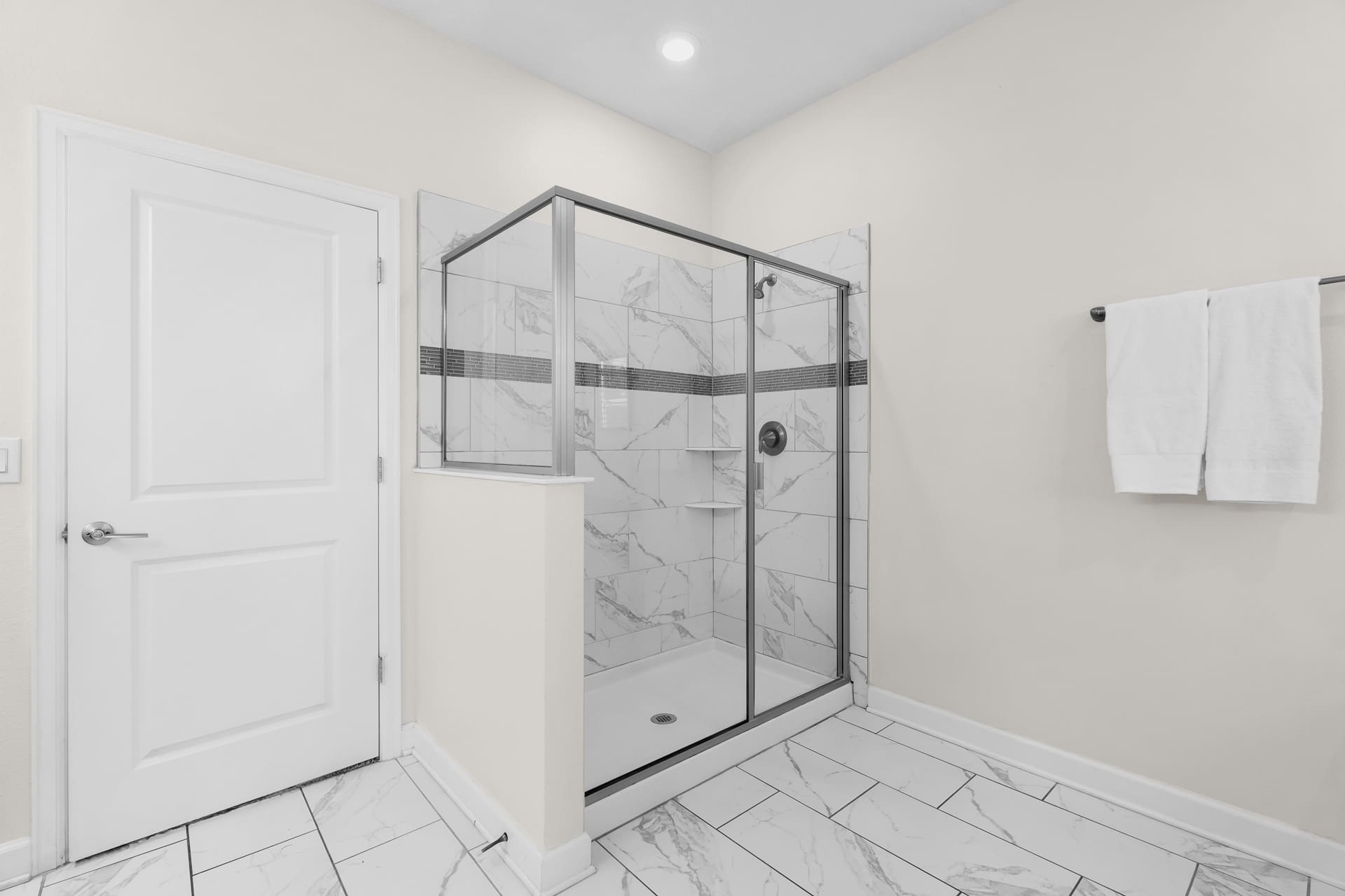 Modern walk-in shower with glass enclosure and white marble-patterned tiles, featuring a dark shower head and a built-in shelf, adjacent to a white door.
