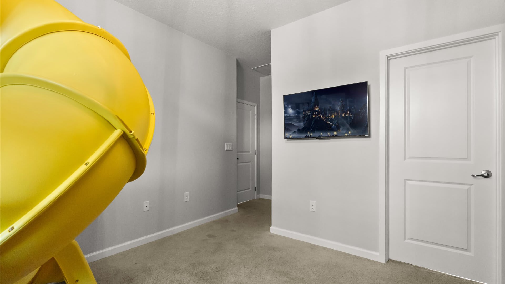 A section of a light-colored carpeted room showcases a large yellow spiral slide, a wall-mounted television displaying a fantasy scene, and two white doors leading to other areas.