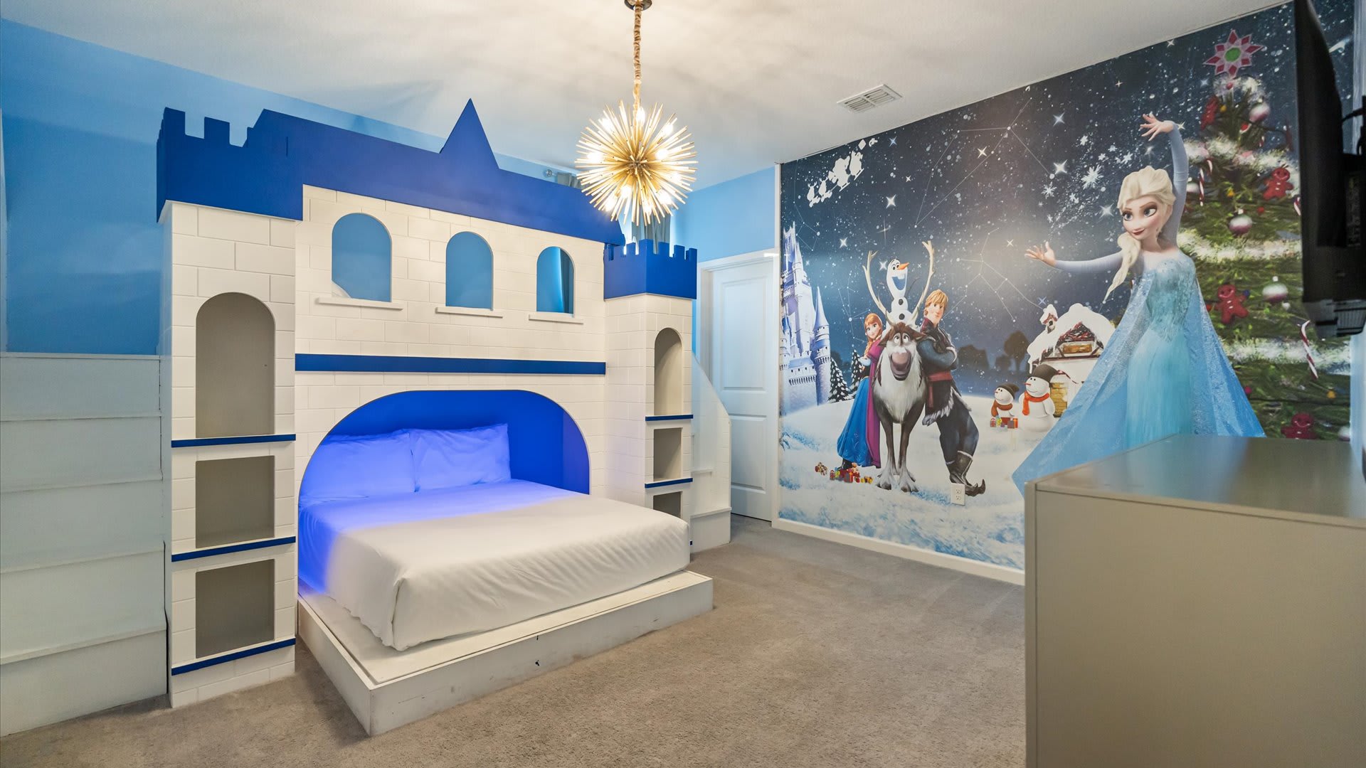 A magical Frozen-themed bedroom featuring a large white and blue castle-shaped bed with a built-in full-size mattress. A vibrant mural of Elsa, Anna, and Olaf in a winter wonderland covers one wall.