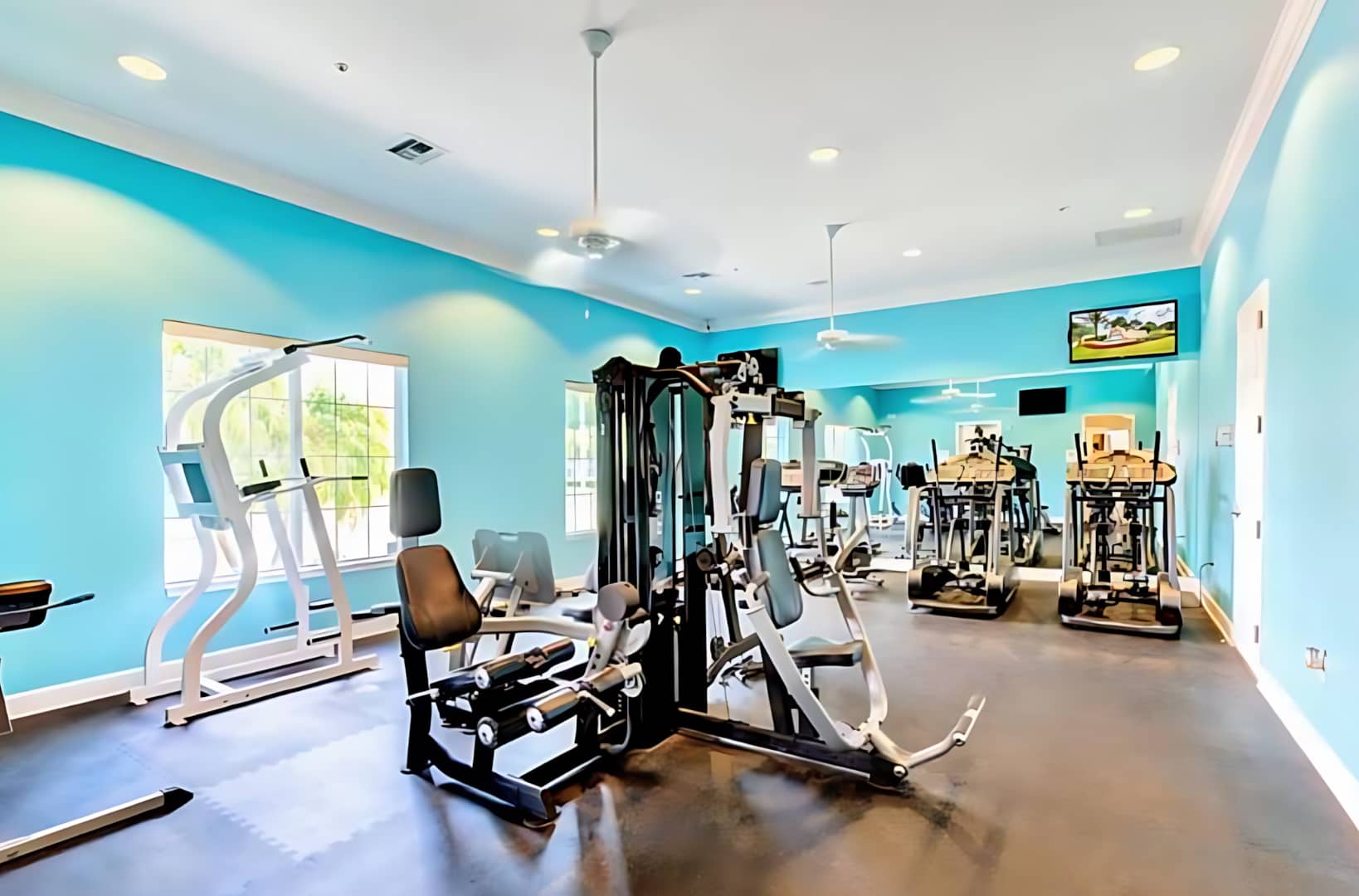 Well-equipped fitness center with bright blue walls and light flooring, featuring various exercise machines including treadmills, ellipticals, and weight machines. Large windows provide natural light.