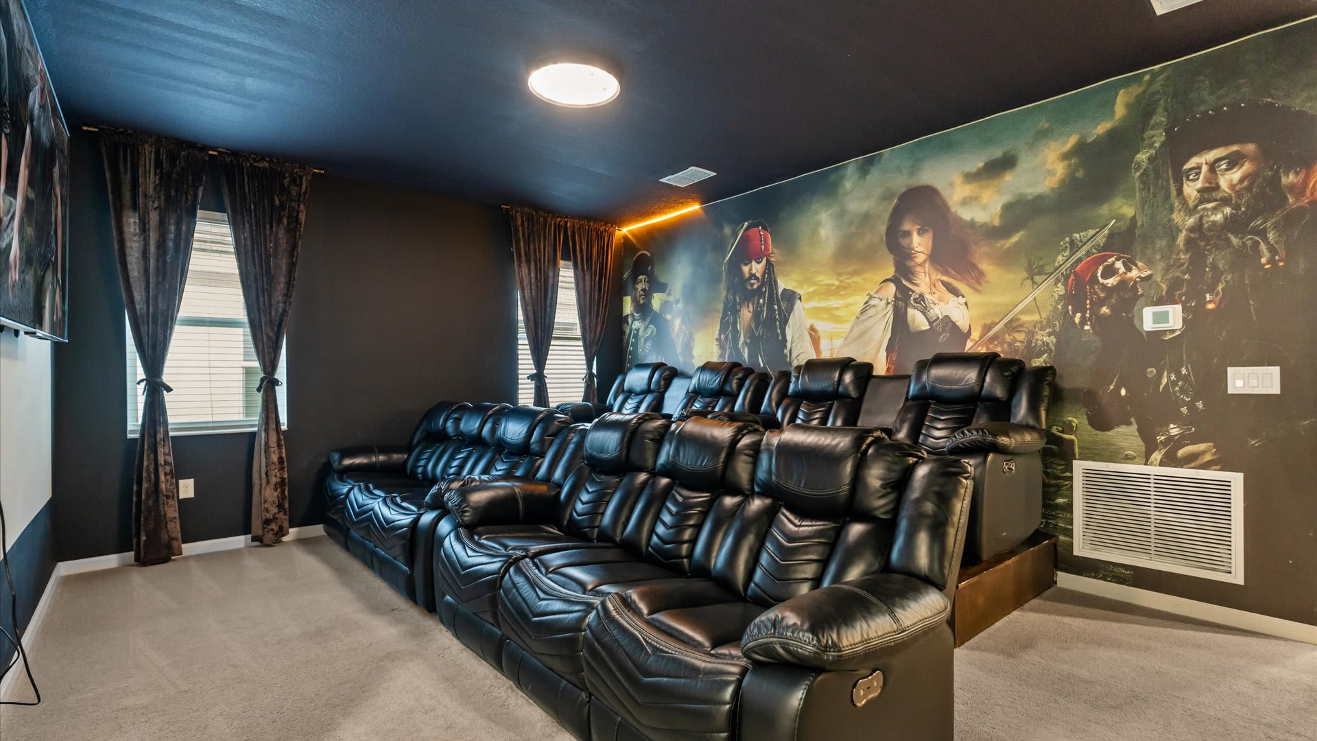 Dark-themed home cinema with multiple rows of plush black reclining seats, featuring a large wall mural of Pirates of the Caribbean characters and a subtle orange accent light.