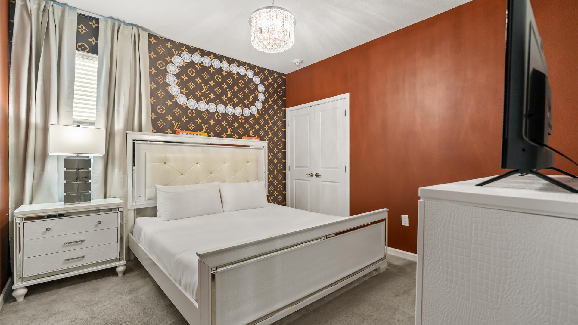 A stylish bedroom features a white upholstered bed with a tufted headboard, a mirrored nightstand, and a unique accent wall with a brown and gold patterned wallpaper and a large oval mirror.