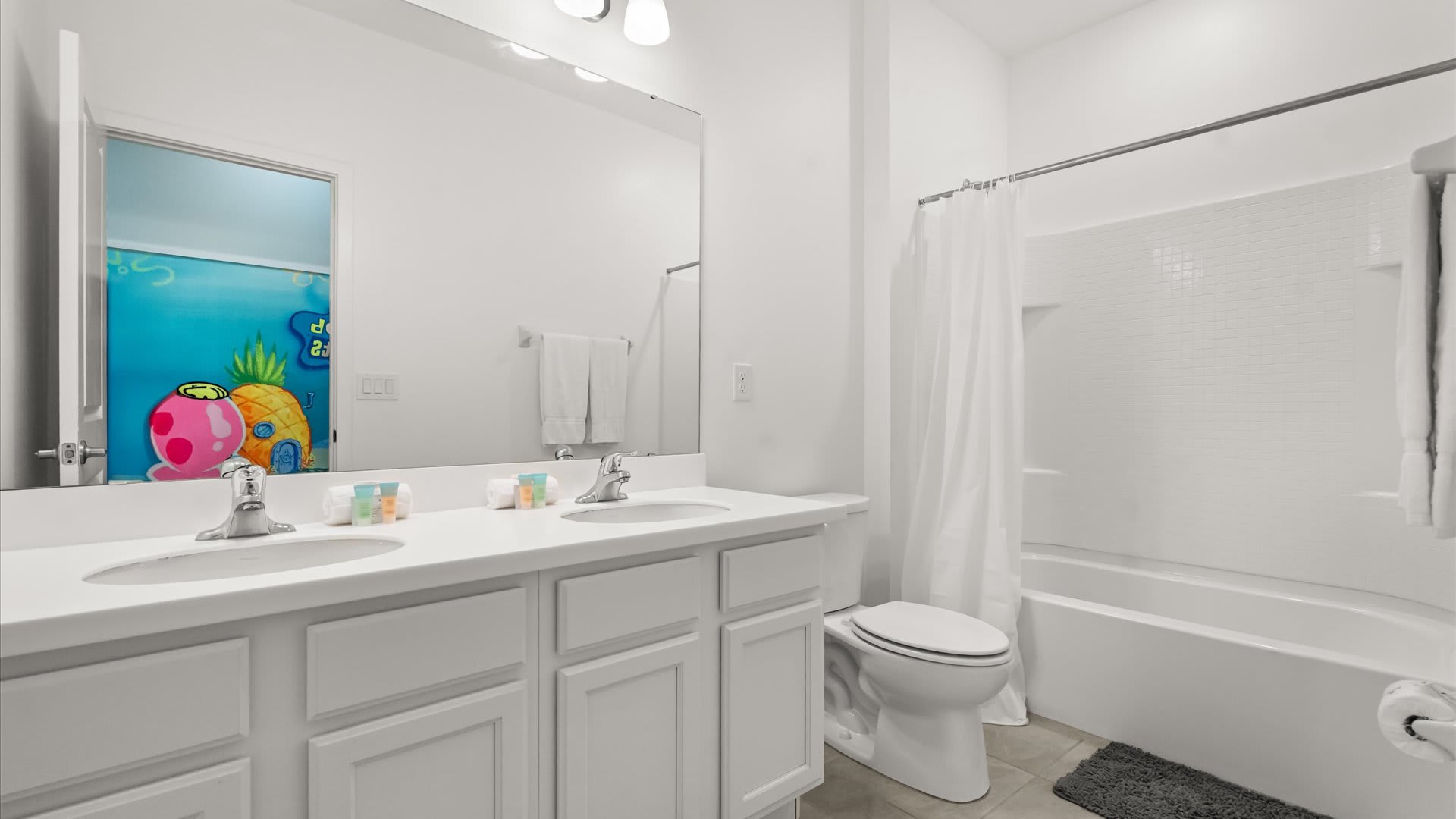 A spacious white bathroom featuring a double vanity with two sinks and a large mirror. A toilet is positioned next to the vanity, and a shower/tub combo with a white curtain is visible.