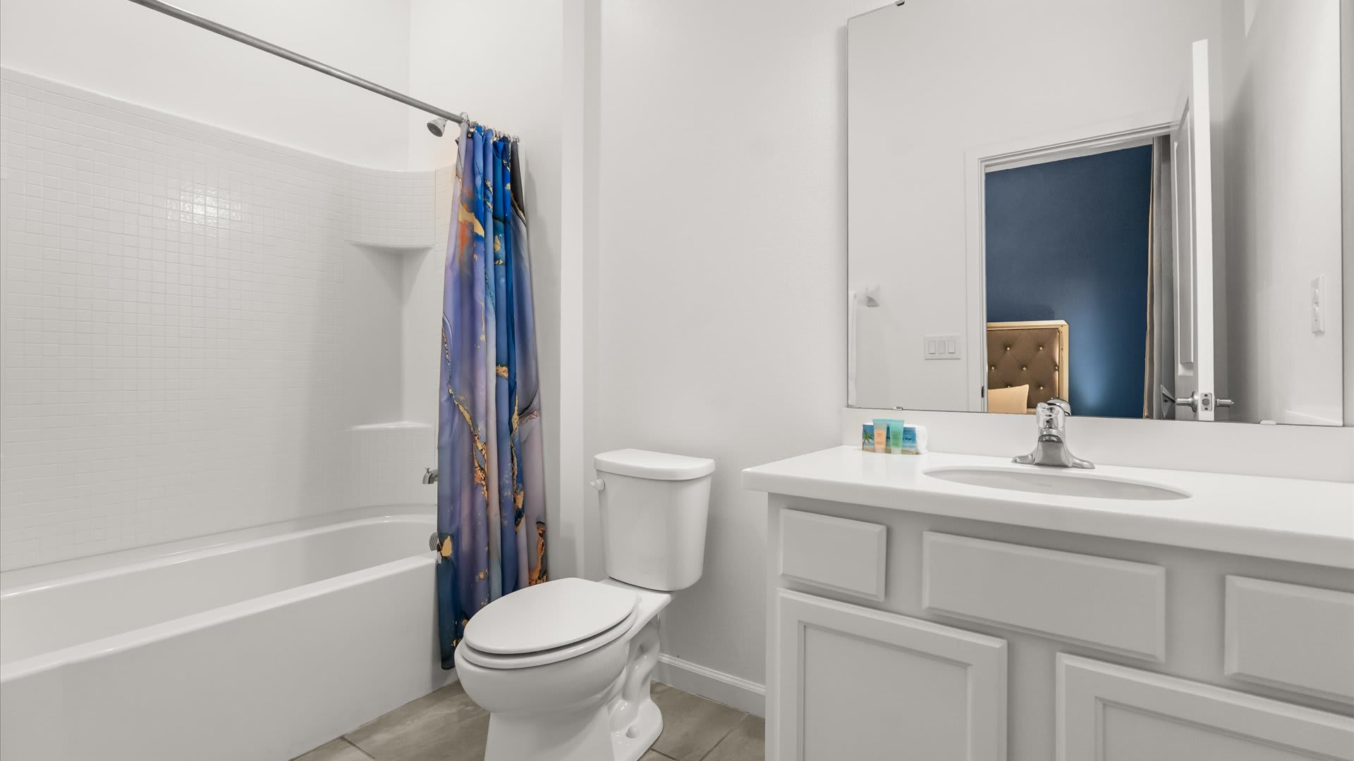 A bright white bathroom featuring a single vanity with a white countertop and sink, a toilet, and a bathtub with a shower curtain displaying a blue and orange abstract design. A large mirror reflects the adjacent bedroom's blue wall.