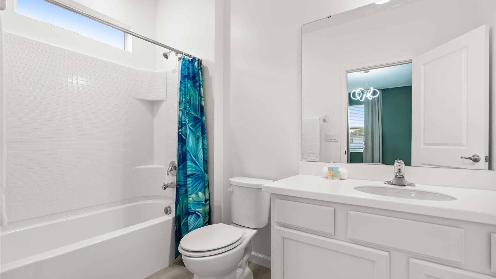 A bright white bathroom featuring a bathtub with a shower and a vibrant blue tropical-patterned shower curtain. It includes a single vanity with a white countertop, a large mirror, and a toilet.