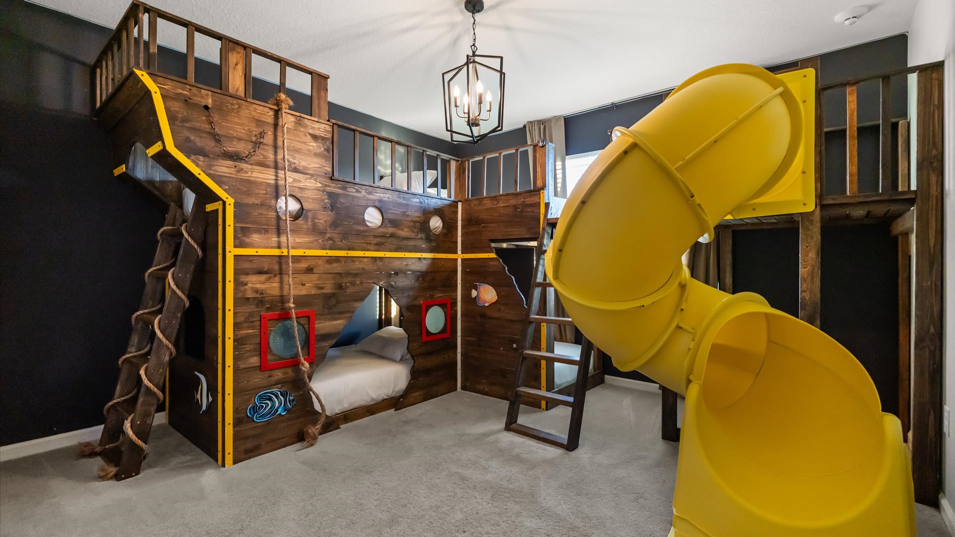 A themed kids' bedroom features an elaborate wooden pirate ship bunk bed structure with a rope ladder, porthole windows, and a bright yellow spiral slide, all set against dark gray walls.