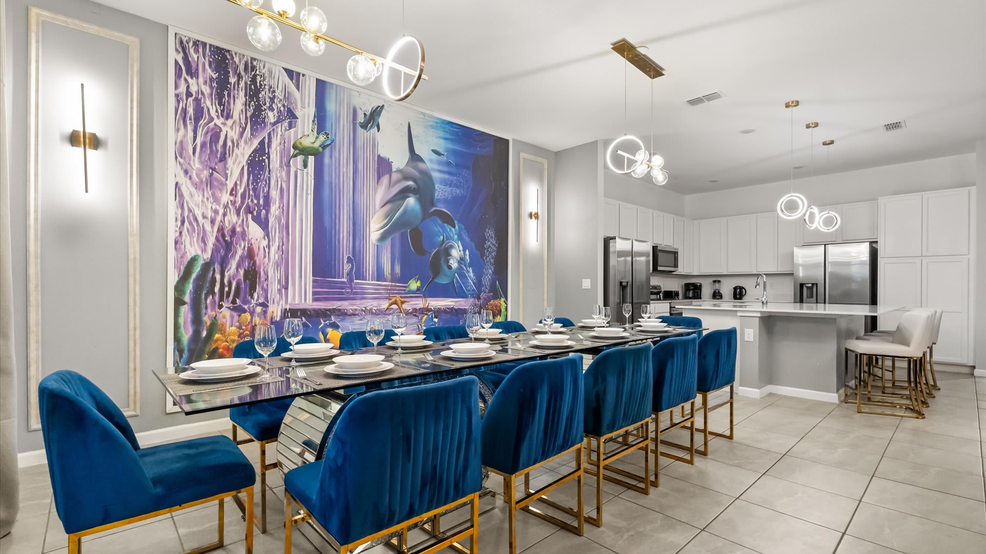 A long, modern dining table with a glass top and gold base is surrounded by royal blue velvet dining chairs, set against a dramatic underwater-themed wall mural, with contemporary pendant lights overhead.