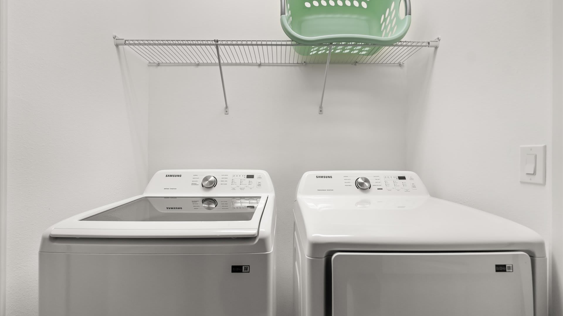 A laundry room is equipped with a white Samsung top-load washing machine and a matching dryer, with a wire shelf mounted above holding a green laundry basket.
