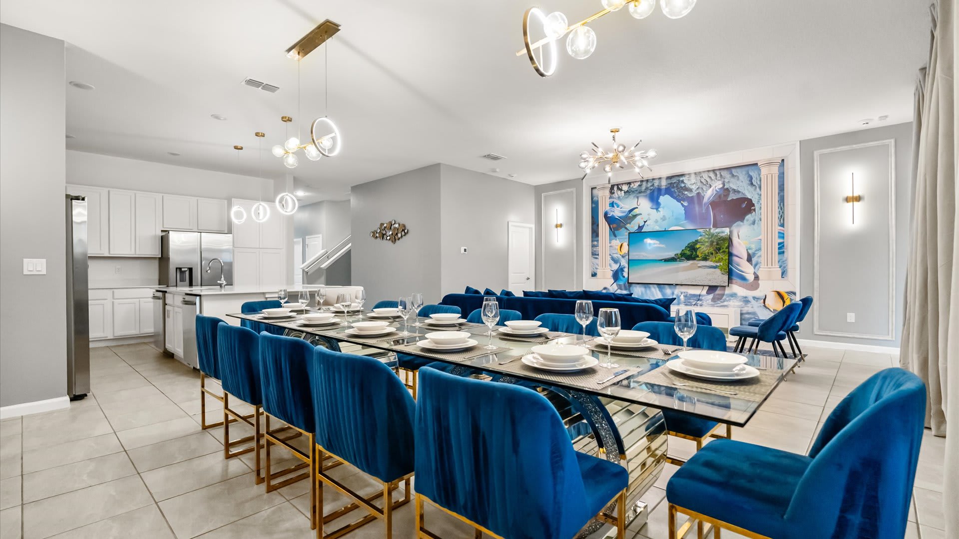 The elegant dining area features a long glass-top table with royal blue velvet chairs, positioned beneath modern circular pendant lights, with an open view to the contemporary white kitchen in the background.