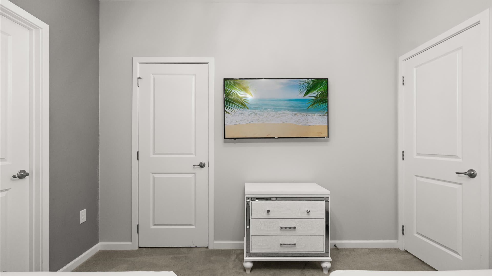 A bright bedroom wall with a large flat-screen TV mounted above a white mirrored dresser featuring three drawers. Two white doors flank the dresser, and the room has light gray carpet.