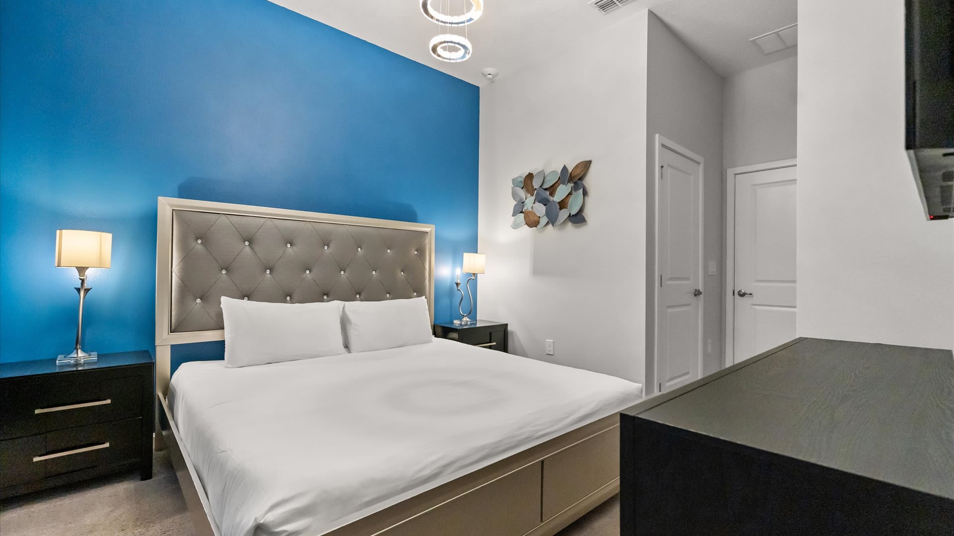 A modern bedroom featuring a king-sized bed with a gray tufted headboard against a vibrant blue accent wall, flanked by dark wood nightstands with silver lamps. Overhead, a contemporary chandelier with multiple rings illuminates the space.