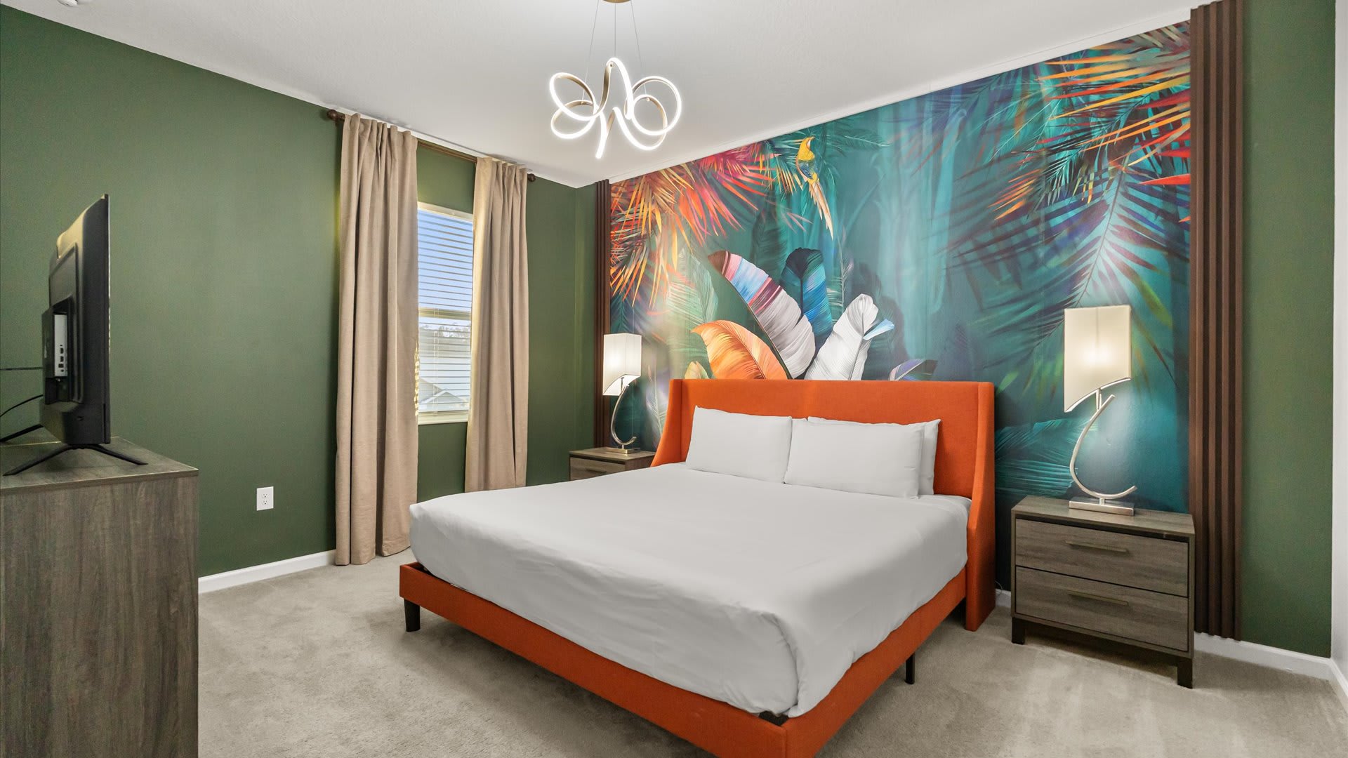 A vibrant bedroom featuring a king-sized bed with an orange frame, flanked by dark nightstands and lamps. A large tropical-themed mural covers the wall behind the bed, contrasting with dark green walls and light curtains.