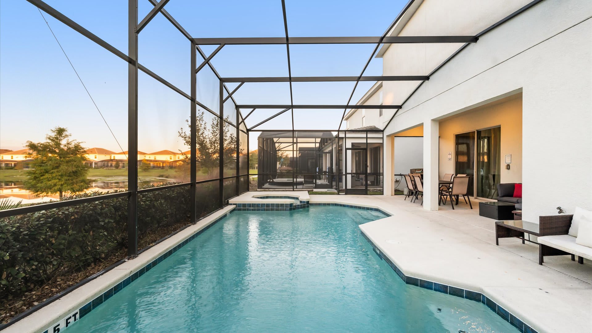 Expansive screened-in private pool area with an integrated spa, surrounded by a light concrete deck and offering serene views of a lake and distant residential homes under a clear sky.