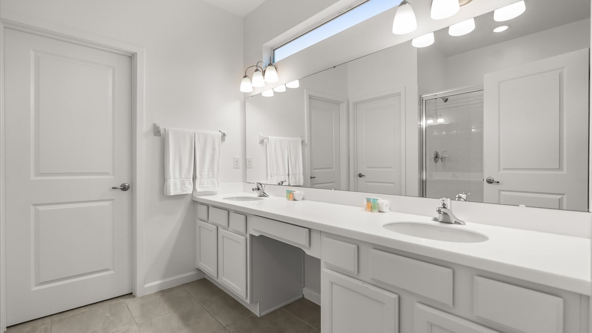 A spacious white bathroom features a long double vanity with two sinks and white countertops, a large mirror, and a separate glass-enclosed shower visible in the reflection, with light-colored floor tiles.