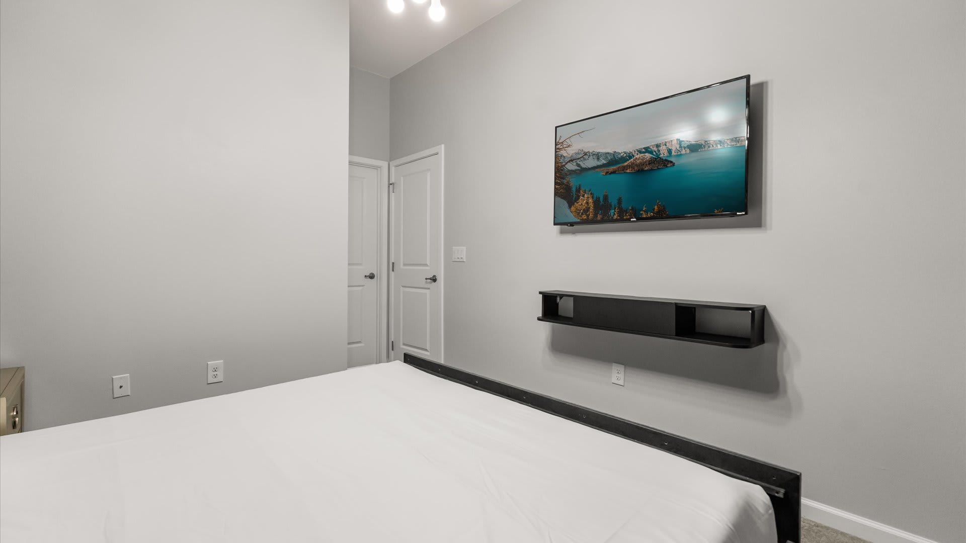The green-themed bedroom features a wall-mounted flat-screen TV displaying a scenic lake view, positioned above a floating black shelf. The room has light gray walls and a simple bed frame with white bedding.