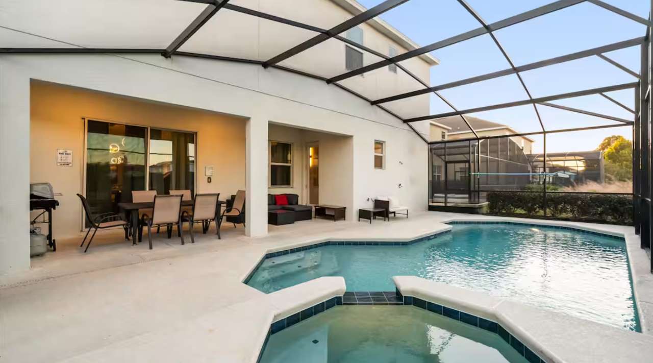 Spacious screened-in patio with a private swimming pool and hot tub, featuring an outdoor dining set and lounge seating, overlooking a landscaped backyard and distant homes.