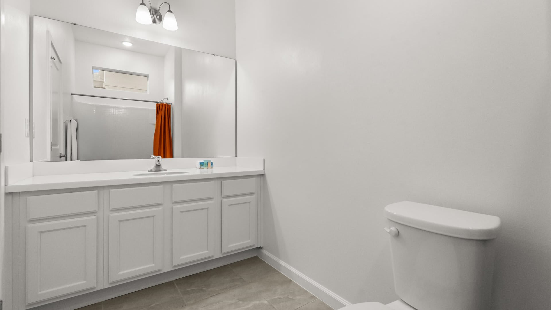 A spacious white bathroom with a long single vanity featuring a white countertop and sink, and a toilet. A large mirror extends across the vanity, reflecting the bright white walls.