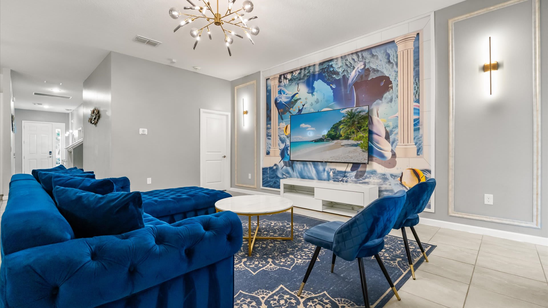 A modern living room features a large wall-mounted TV above a white media console, set against a vibrant blue and white wall mural, complemented by a royal blue velvet sectional sofa and two matching accent chairs.