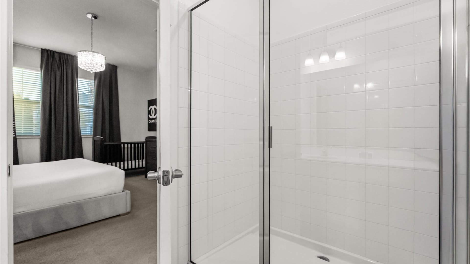 A modern shower enclosure with clear glass doors and white subway tiles. The view extends into the adjacent bedroom, showing a bed and a crib.