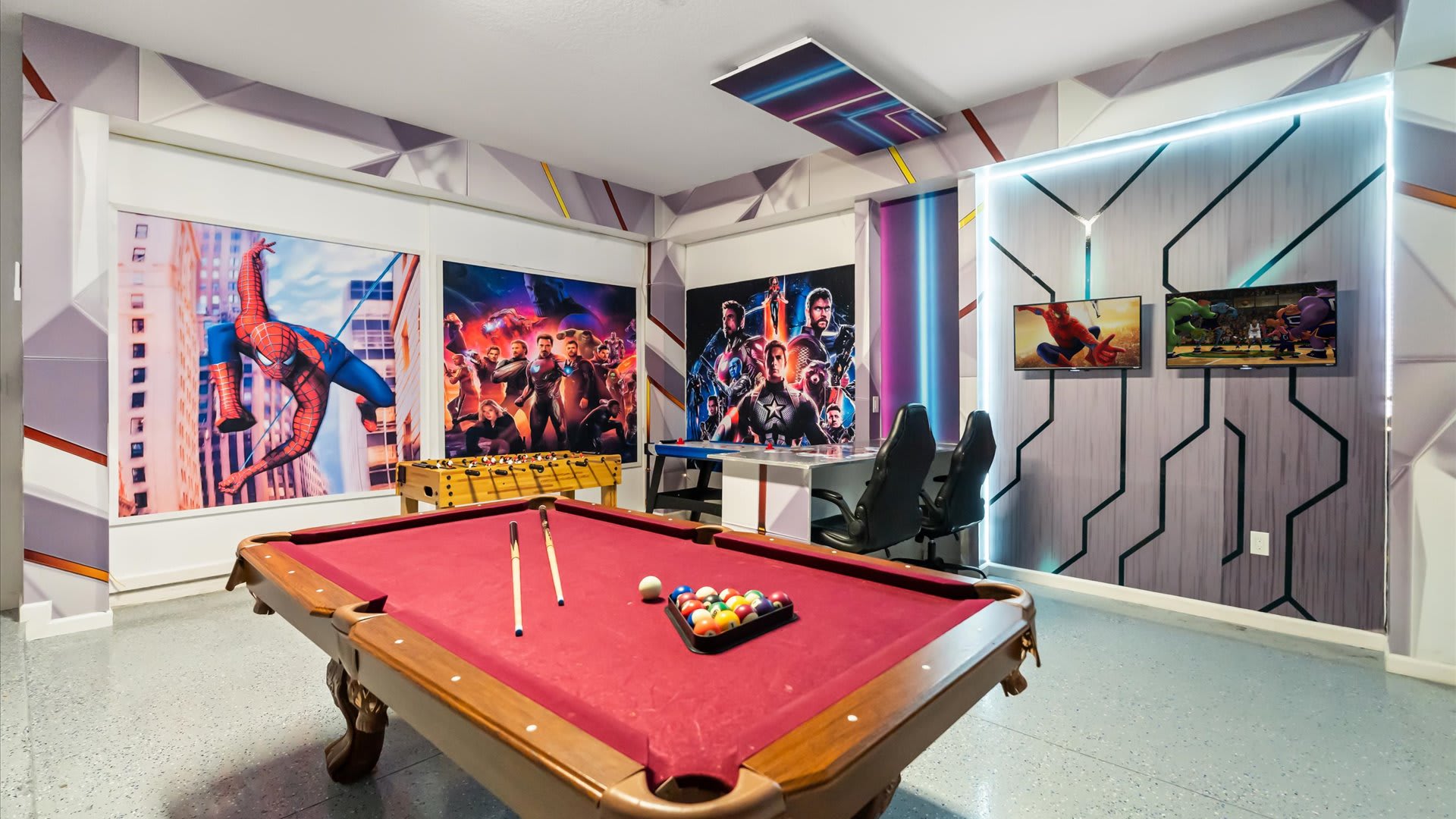 Modern game room with a red-felt pool table and foosball, adorned with geometric wall patterns and large superhero-themed murals, featuring multiple flat-screen TVs and gaming chairs.