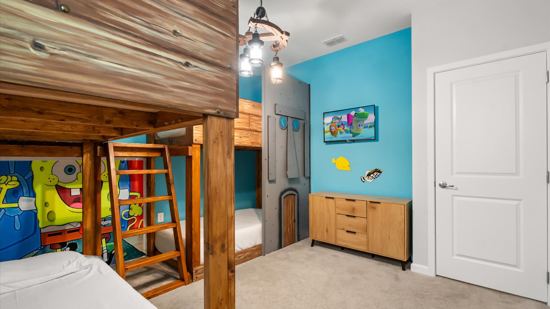 Another view of the Spongebob themed bedroom, showing a flat-screen TV mounted on a blue wall above a light wood dresser. A custom Spongebob mural and a gray themed wall panel resembling a submarine are also visible.