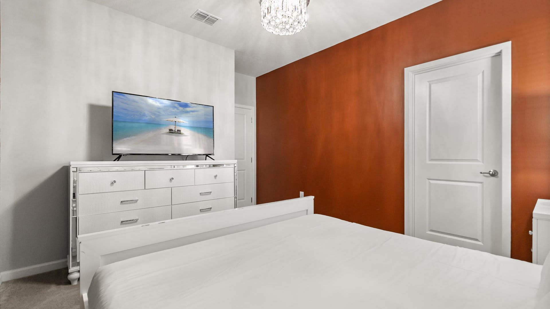 This bedroom includes a white dresser with mirrored accents and a flat-screen TV, positioned opposite a white bed, with a warm rust-colored accent wall and a white door visible.