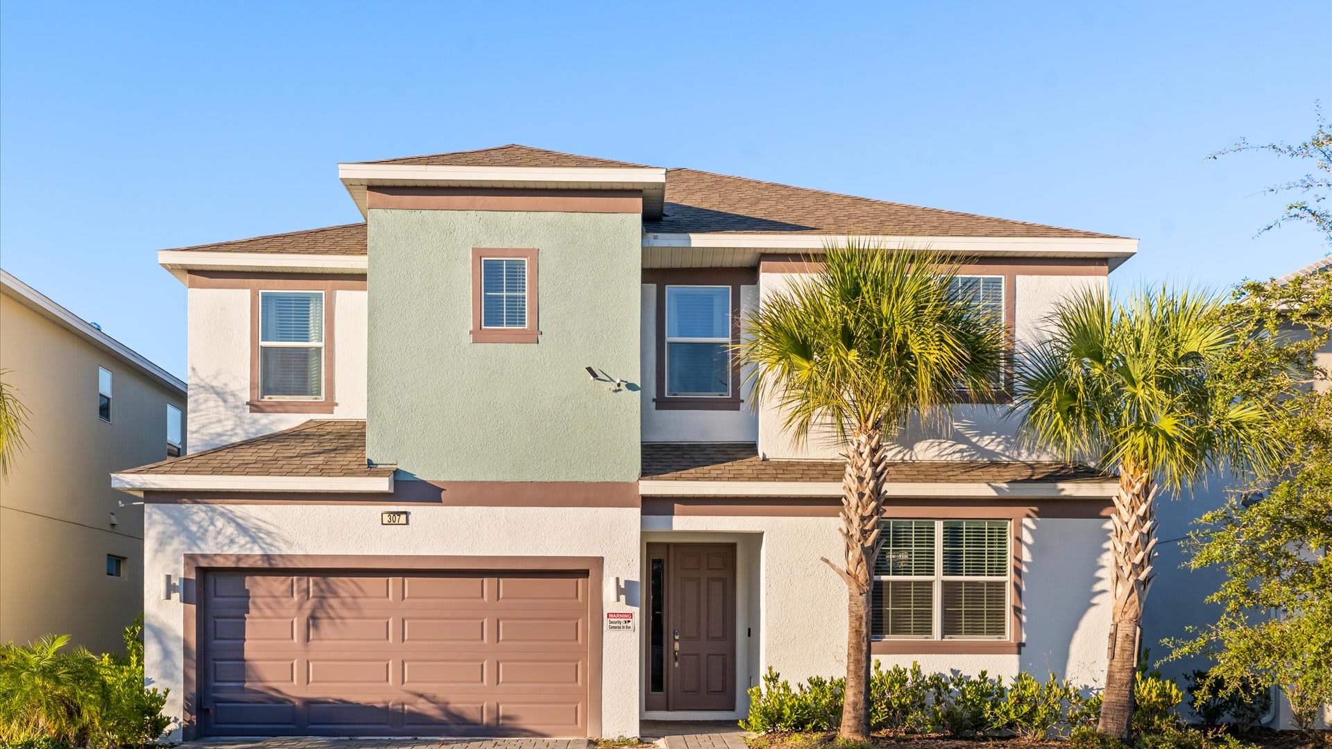 A two-story house with a light-colored stucco exterior, brown trim, and a central section painted light green, features a brown garage door, a front door, and two palm trees in the front yard under a clear blue sky.
