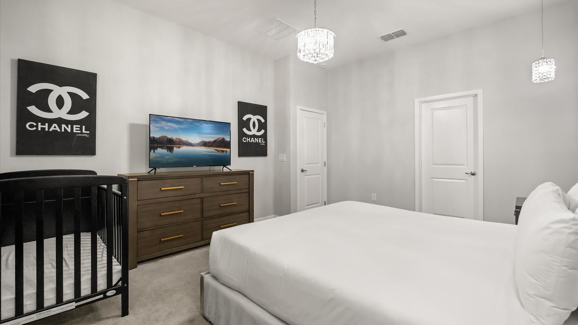 Another view of the bedroom, showing a large flat-screen TV above a wooden dresser with gold handles. Two "Chanel" themed art pieces adorn the light grey walls, and a black crib is positioned next to the bed.