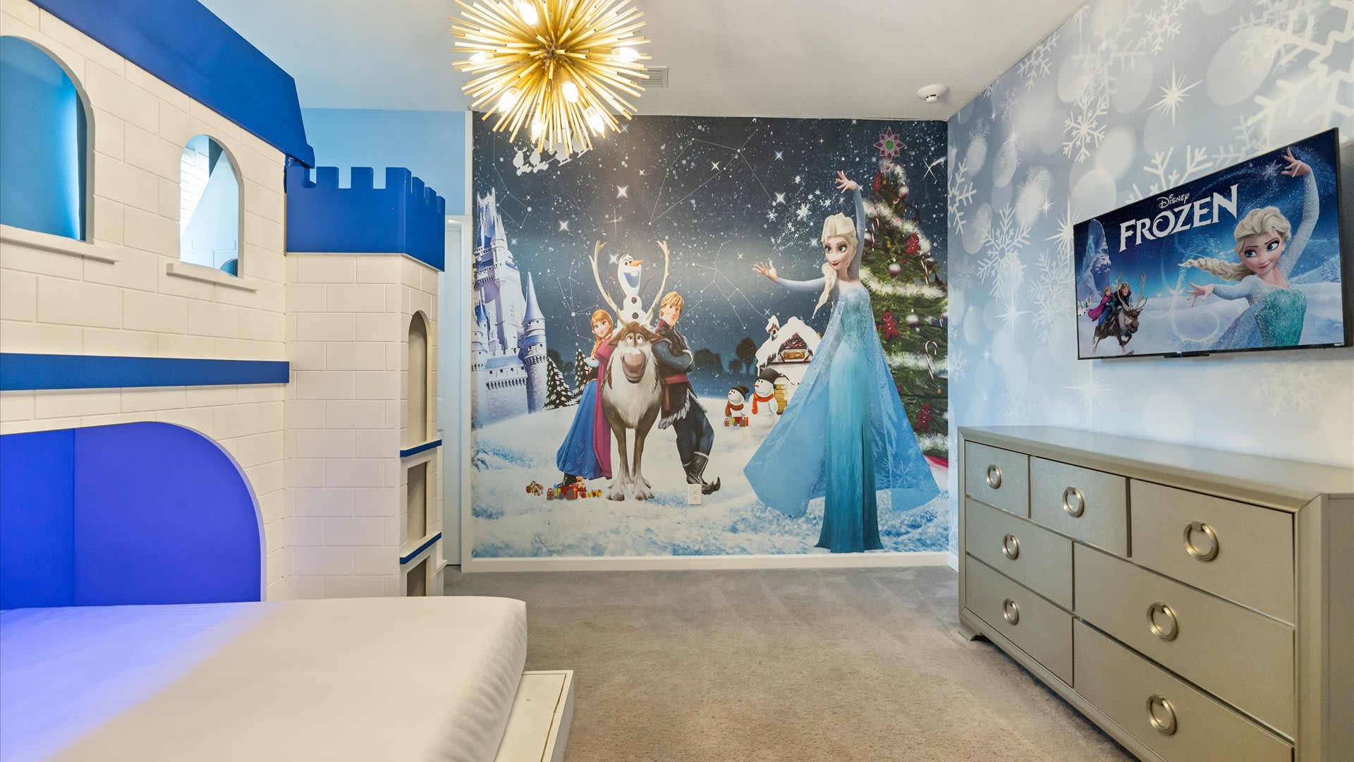 A wide shot of the Frozen-themed bedroom, showcasing the full wall mural of Elsa, Anna, and Olaf in a winter wonderland. A flat-screen TV is mounted on the wall above a gray dresser.
