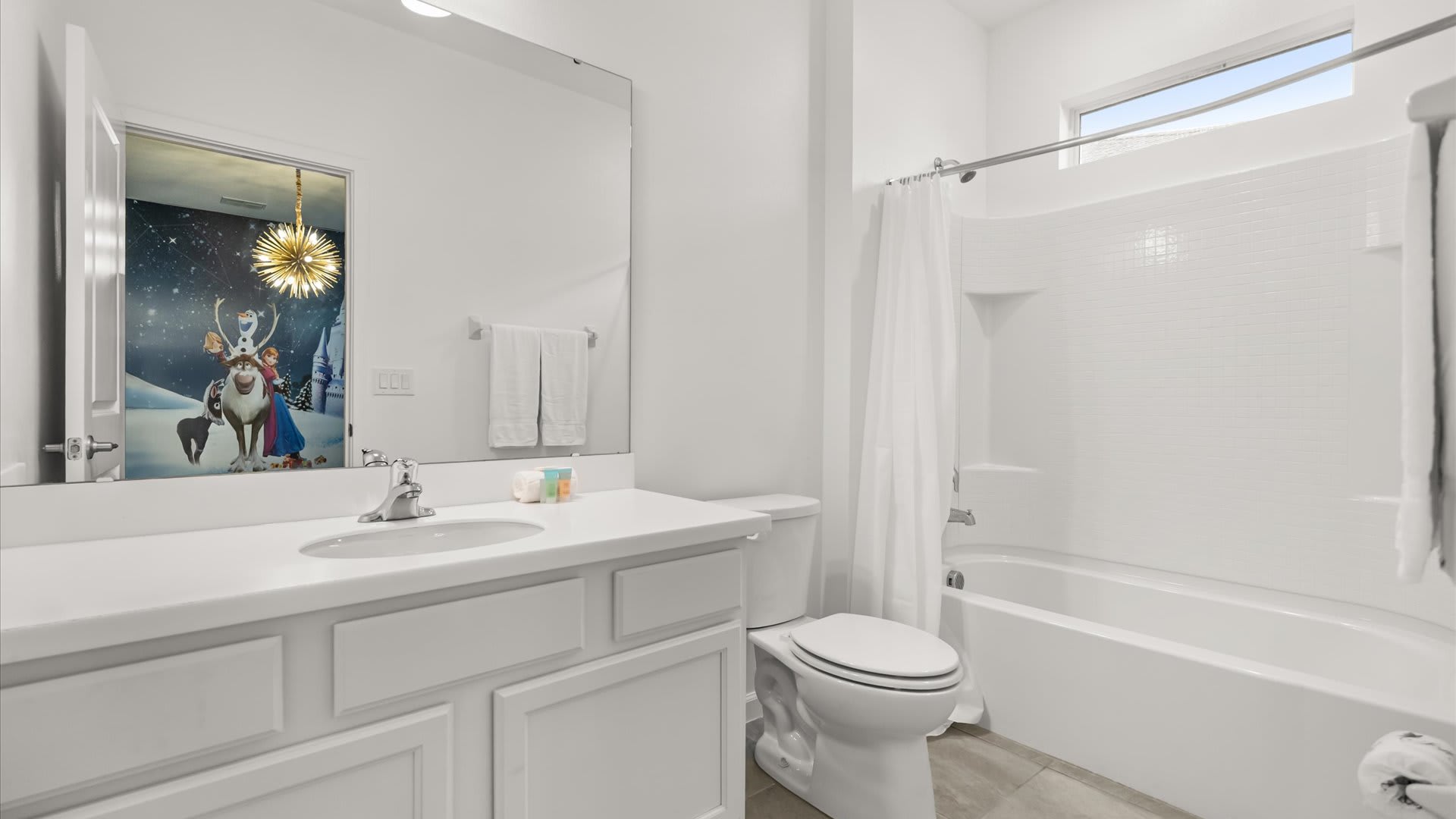 A bright white bathroom features a single vanity with a white countertop and sink, a large mirror reflecting a themed bedroom door, and a white tiled shower/tub combo with a shower curtain.
