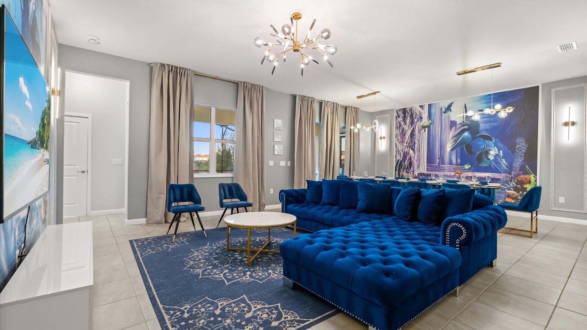 The spacious living room showcases a royal blue velvet sectional and two accent chairs on a patterned blue rug, with light-colored curtains framing a window and a large wall mural visible in the background.
