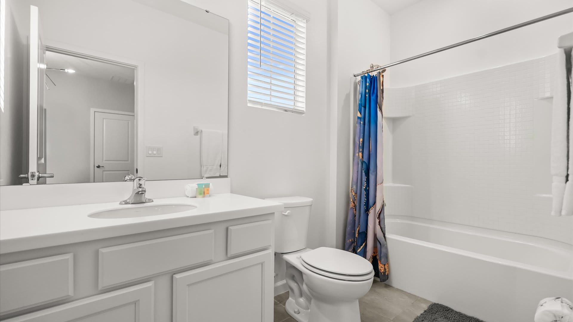 A clean, modern bathroom with a white vanity featuring a single sink and a large mirror. A toilet is next to the vanity, and a shower/tub combo with a blue and white patterned shower curtain is visible.