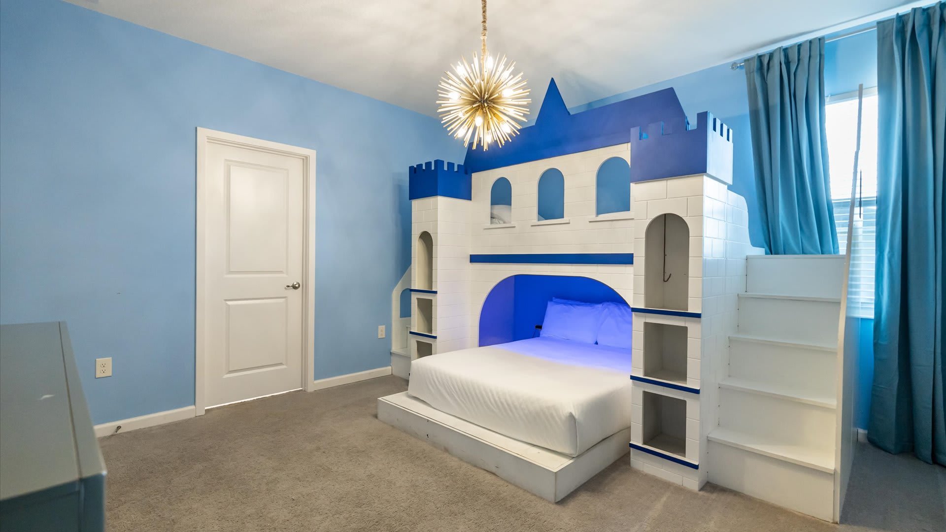 A different angle of the Frozen-themed bedroom, highlighting the impressive white and blue castle-shaped bed with integrated steps and shelving. A white door and a window with blue curtains are visible.
