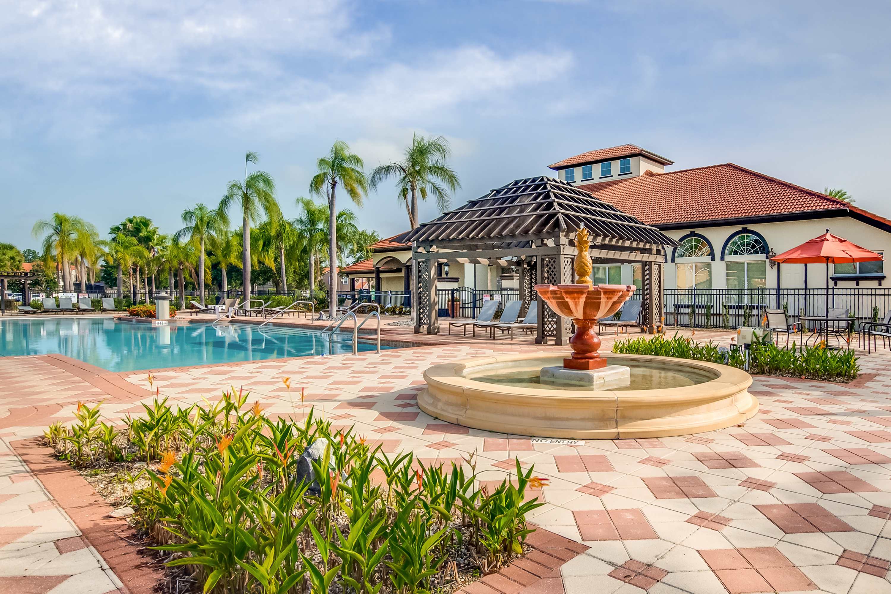Expansive communal pool area with a decorative fountain in the foreground, surrounded by patterned paver decking, numerous lounge chairs, and tall palm trees. The resort clubhouse is visible in the background.