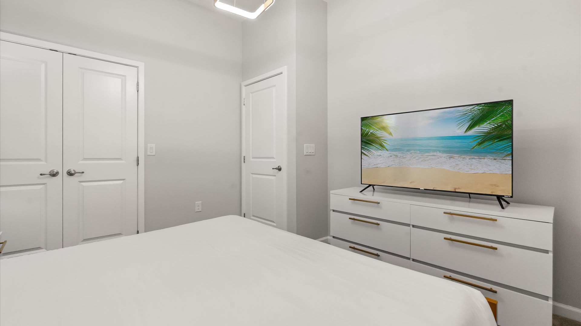 The orange-accented bedroom includes a large white dresser with gold pull handles, topped with a flat-screen TV displaying a beach scene. The room has light gray walls and white doors.