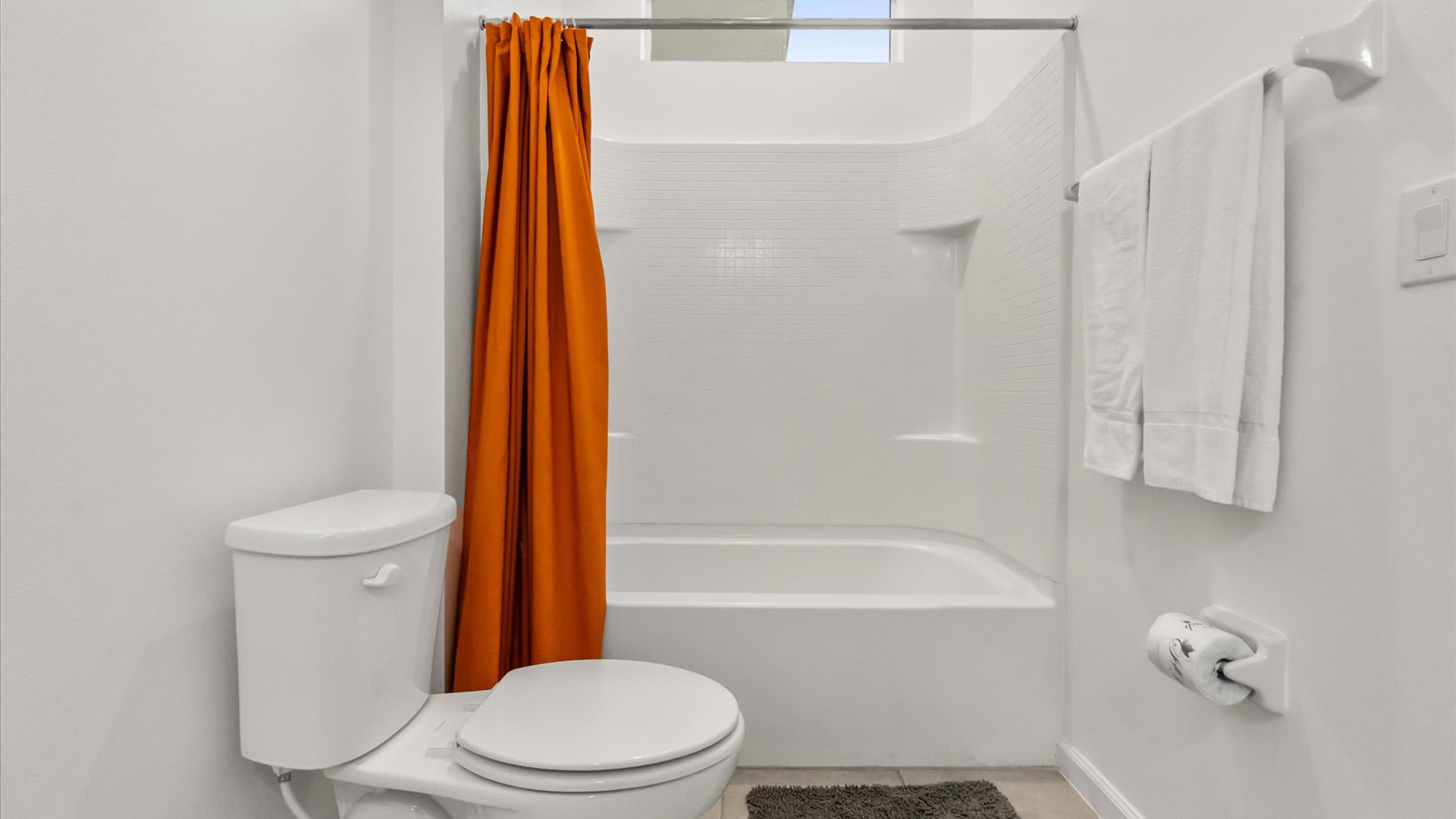 The white bathroom features a bathtub with a bright orange shower curtain, complementing the orange accent wall seen in the associated bedroom. White subway tiles line the shower area, and a gray bath mat is on the floor.