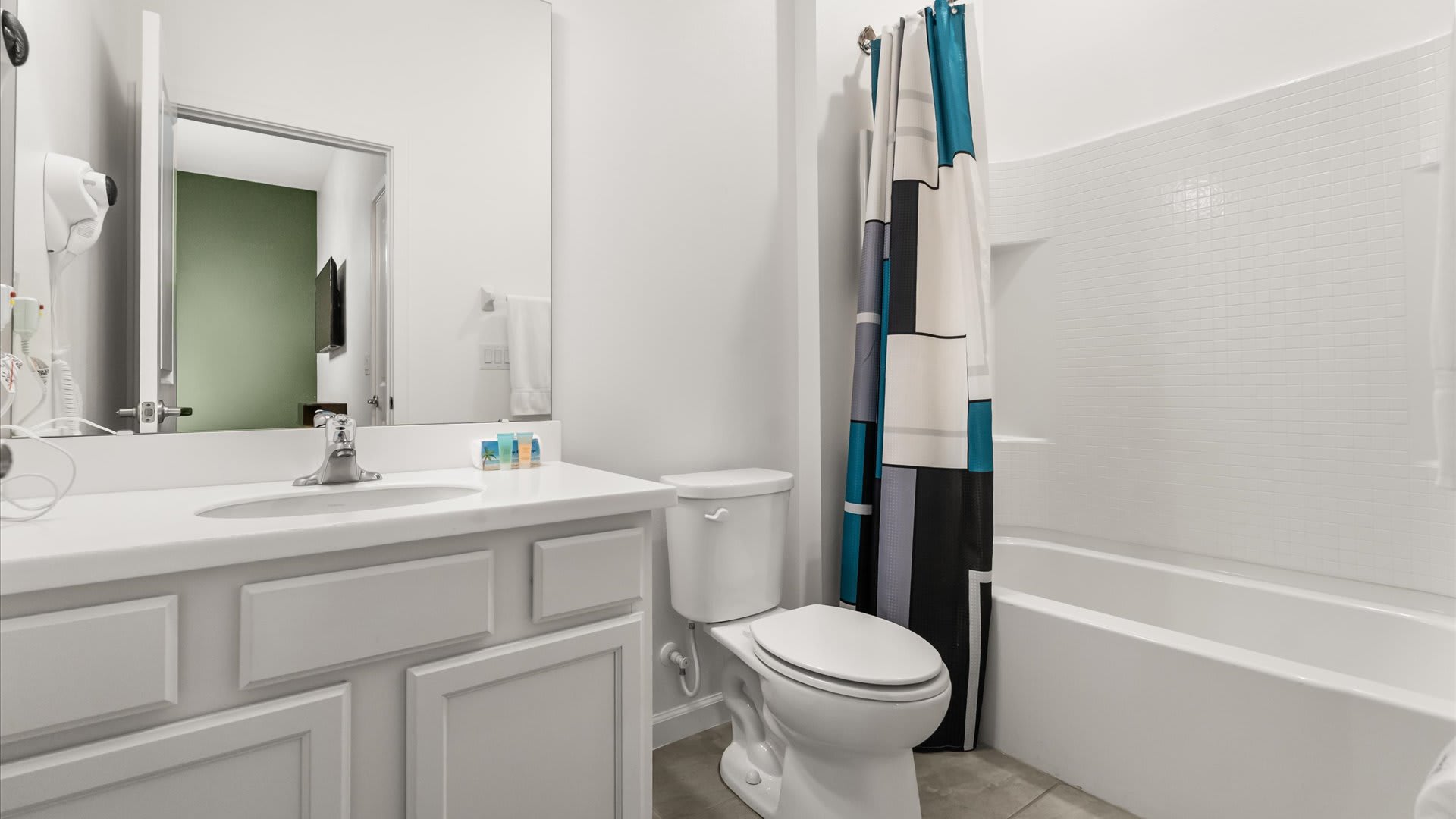 A clean white bathroom with a single vanity, a toilet, and a bathtub featuring a modern geometric shower curtain in blue, gray, and white tones. The mirror reflects a glimpse of the green bedroom wall.