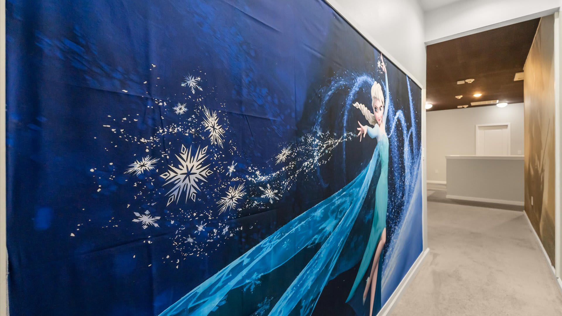 A hallway wall is adorned with a vibrant 'Frozen' themed mural featuring Elsa casting ice magic, set against a dark blue background with sparkling snowflakes, leading to another hallway with a dark ceiling.