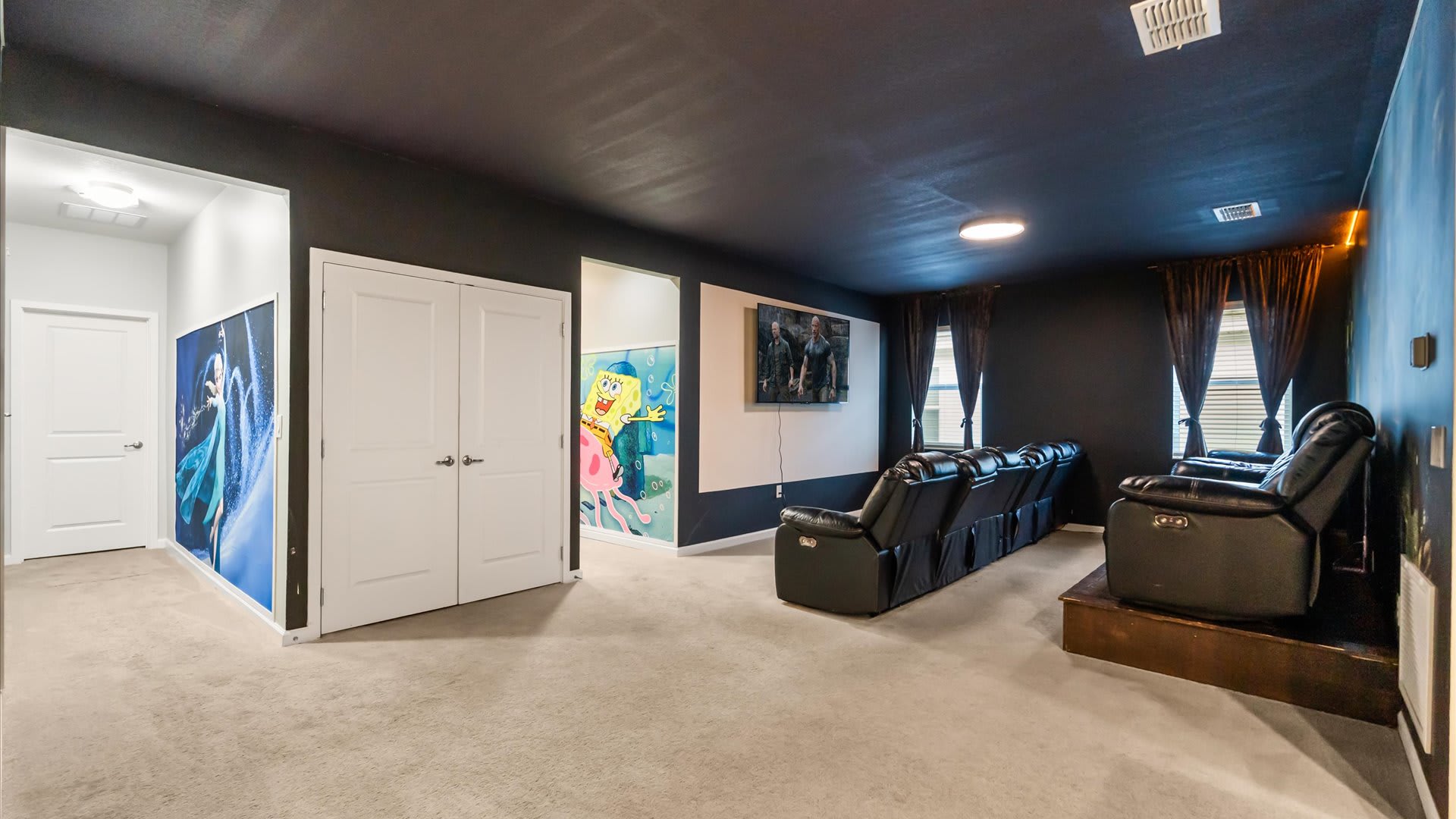Large entertainment room with multiple rows of black reclining theater seats facing a wall-mounted TV, featuring themed wall art of Frozen and Spongebob near the entrance.
