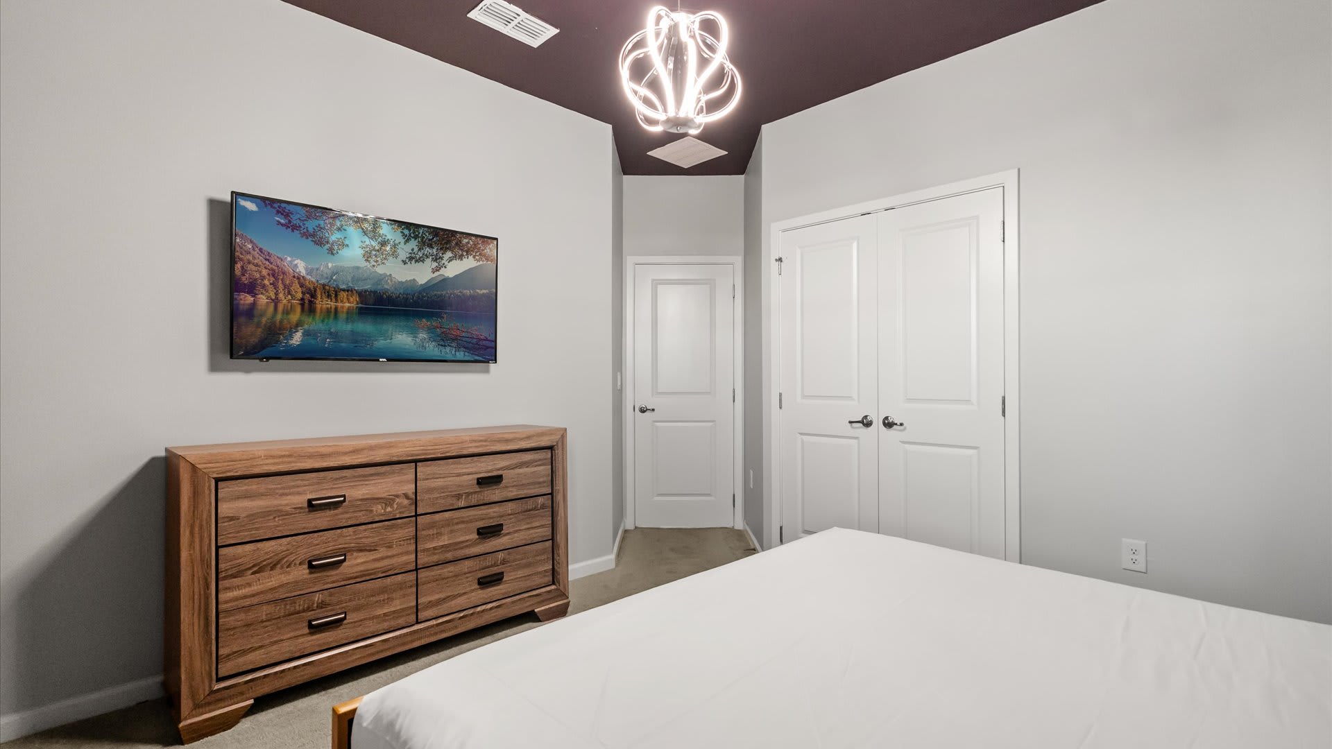 Another view of the bedroom, showing a wall-mounted flat-screen TV above a wooden dresser. The room features light grey walls, white doors, and a distinctive dark ceiling, with a modern chandelier.