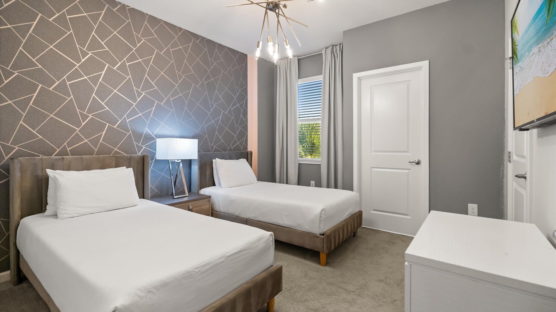 A modern bedroom featuring two twin beds with upholstered headboards, geometric patterned wallpaper on one accent wall, and a large window with gray curtains. A contemporary chandelier hangs from the ceiling, illuminating the carpeted floor.
