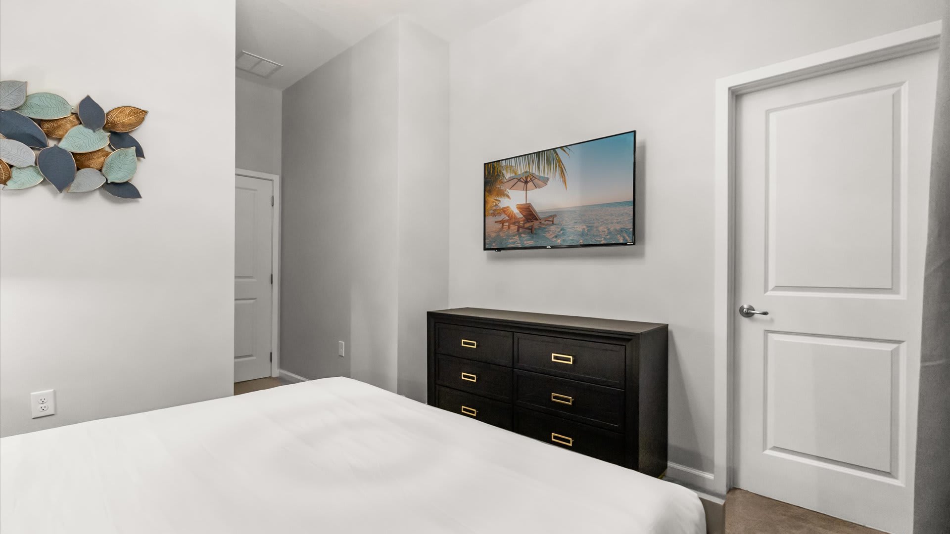The modern bedroom continues with light gray walls, a wall-mounted flat-screen TV displaying a beach scene, and a dark wood dresser with gold pull handles. A decorative leaf-motif wall art piece is visible on the left.
