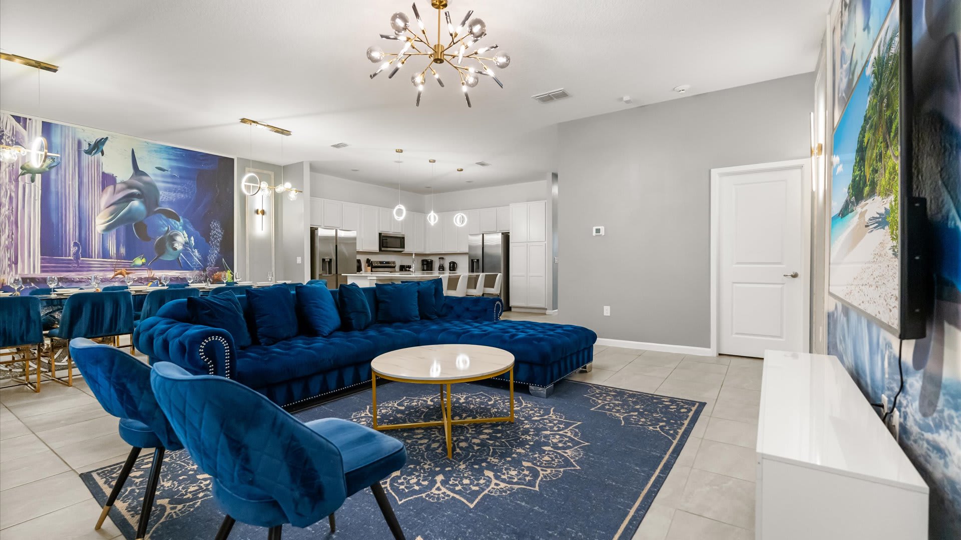 An open-concept living room features a large royal blue velvet sectional sofa, a round gold and marble coffee table, and a modern sputnik chandelier, with a vibrant underwater-themed wall mural extending into the dining area.