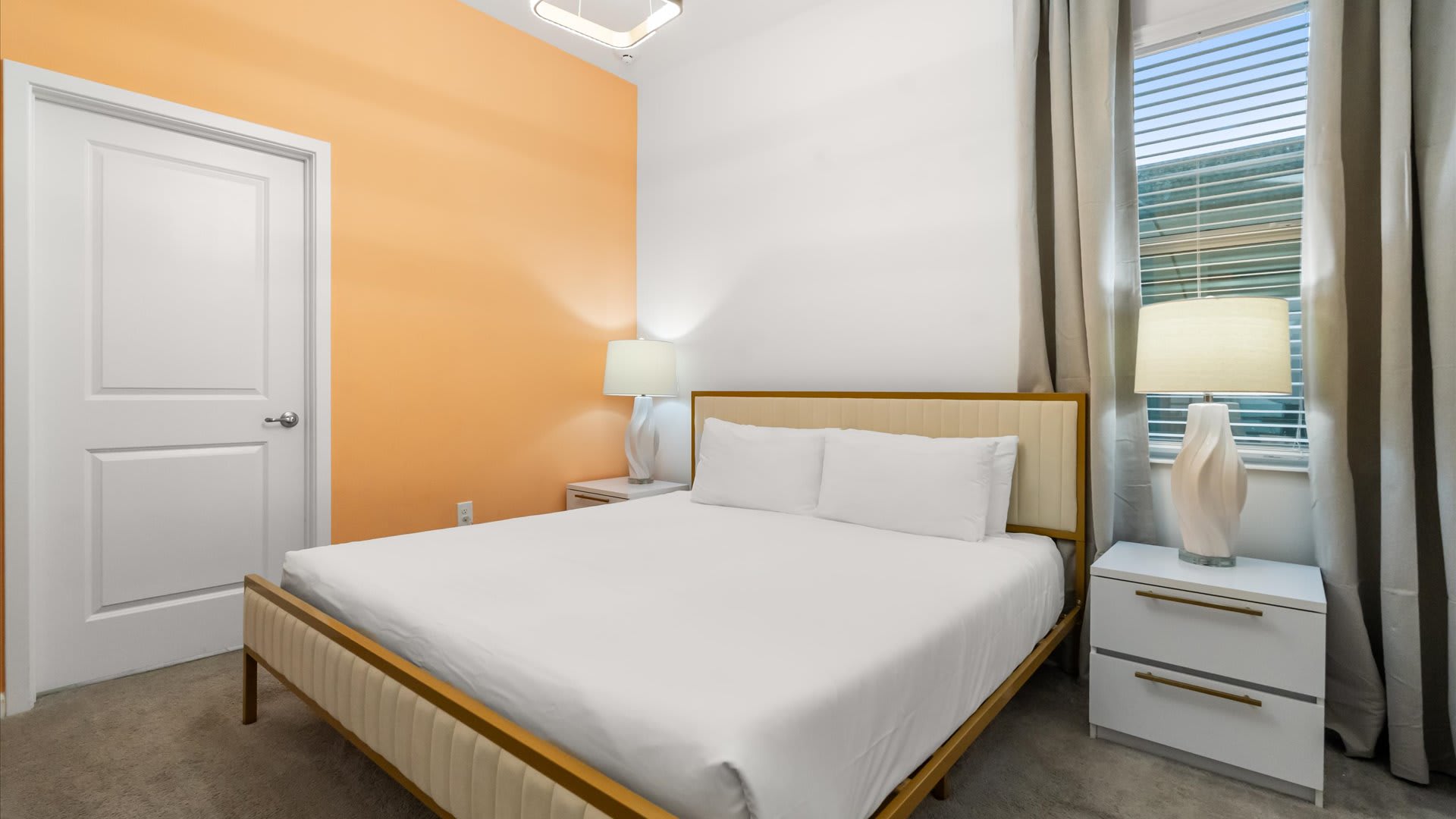 A bright bedroom featuring a queen-sized bed with a gold and cream upholstered headboard against a warm orange accent wall. White nightstands with elegant white lamps flank the bed, and a window with blinds is visible on the right.