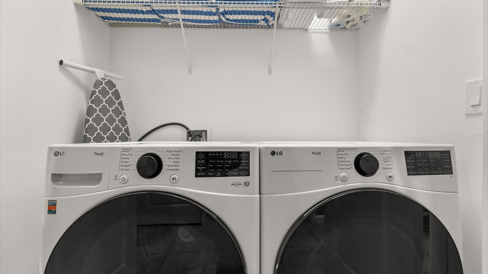This laundry room features a pair of white LG front-load washing machine and dryer, with an ironing board and a wire shelf mounted on the wall above them.