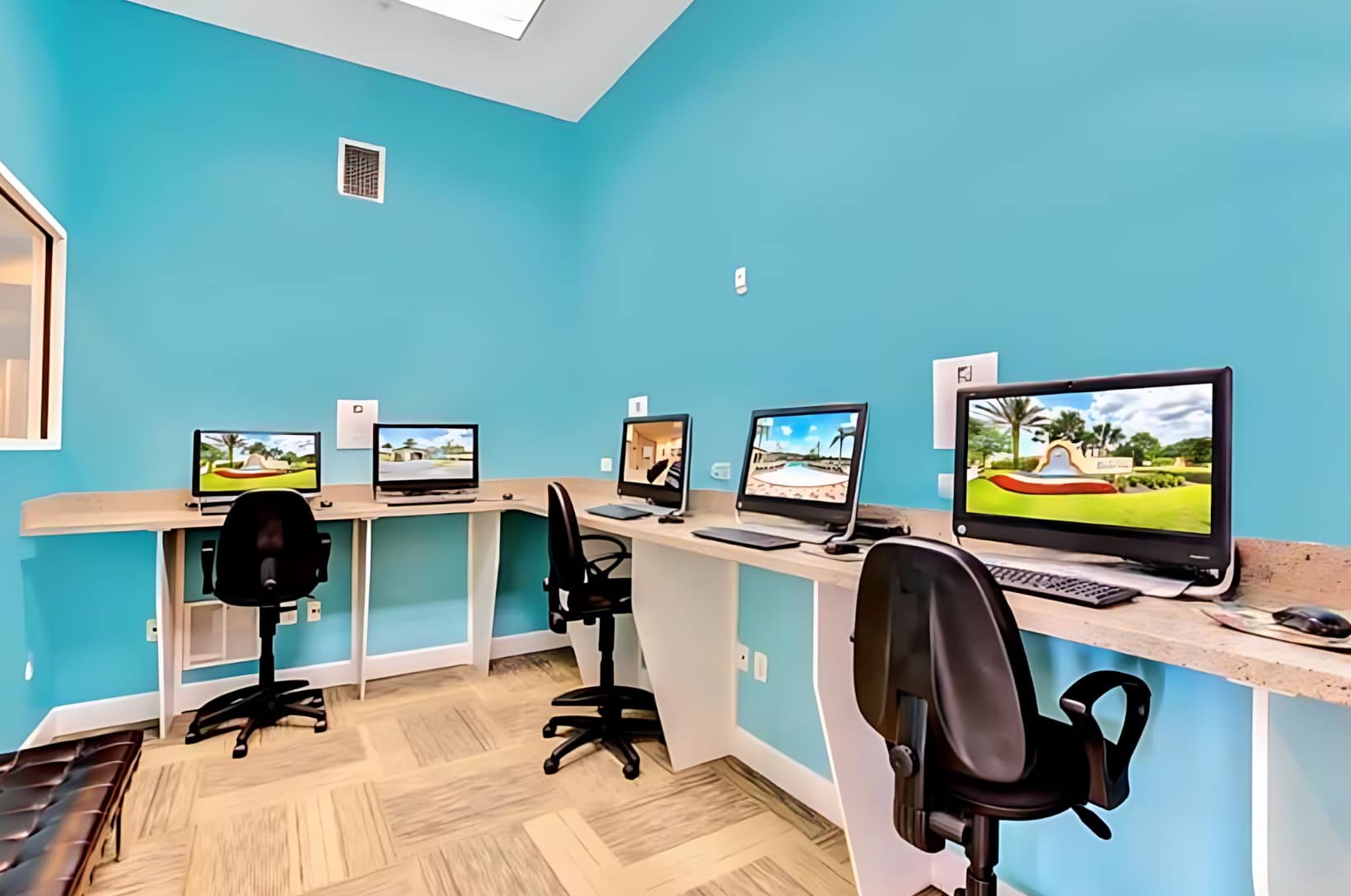 Brightly lit business center with vibrant blue walls, featuring multiple computer workstations with black office chairs and large monitors displaying scenic images. The room has light-colored flooring and a window.