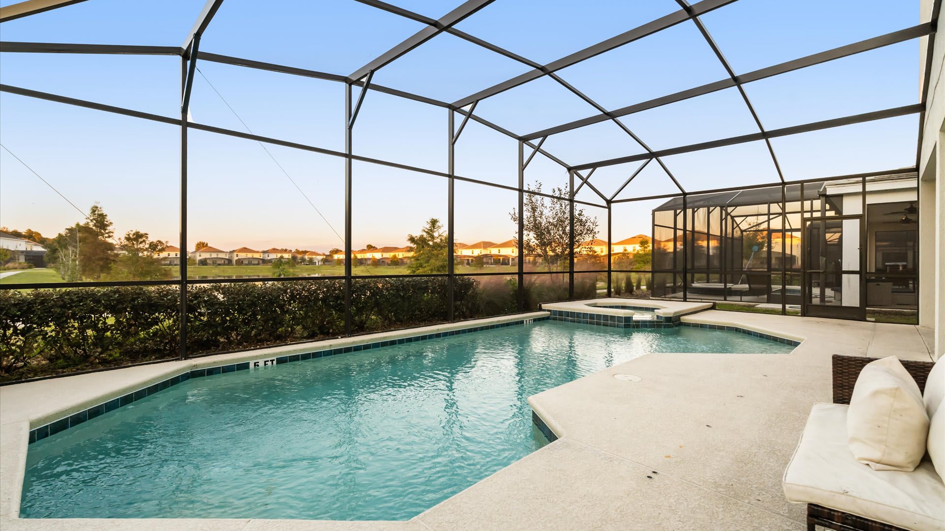 Screened-in private swimming pool with an integrated hot tub, surrounded by a light-colored deck and offering views of a residential area and lush landscaping under a clear sky.
