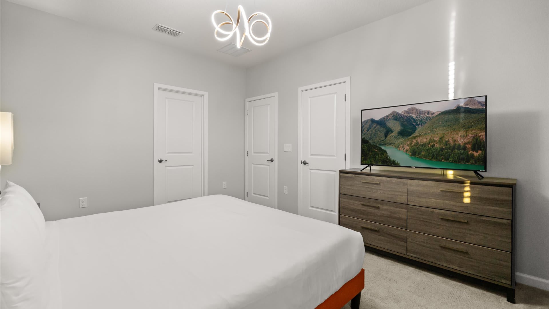 View of the opposite wall in the bedroom, showing a large flat-screen TV mounted above a wooden dresser. The room has light grey walls, white doors, and a modern chandelier hanging from the ceiling.